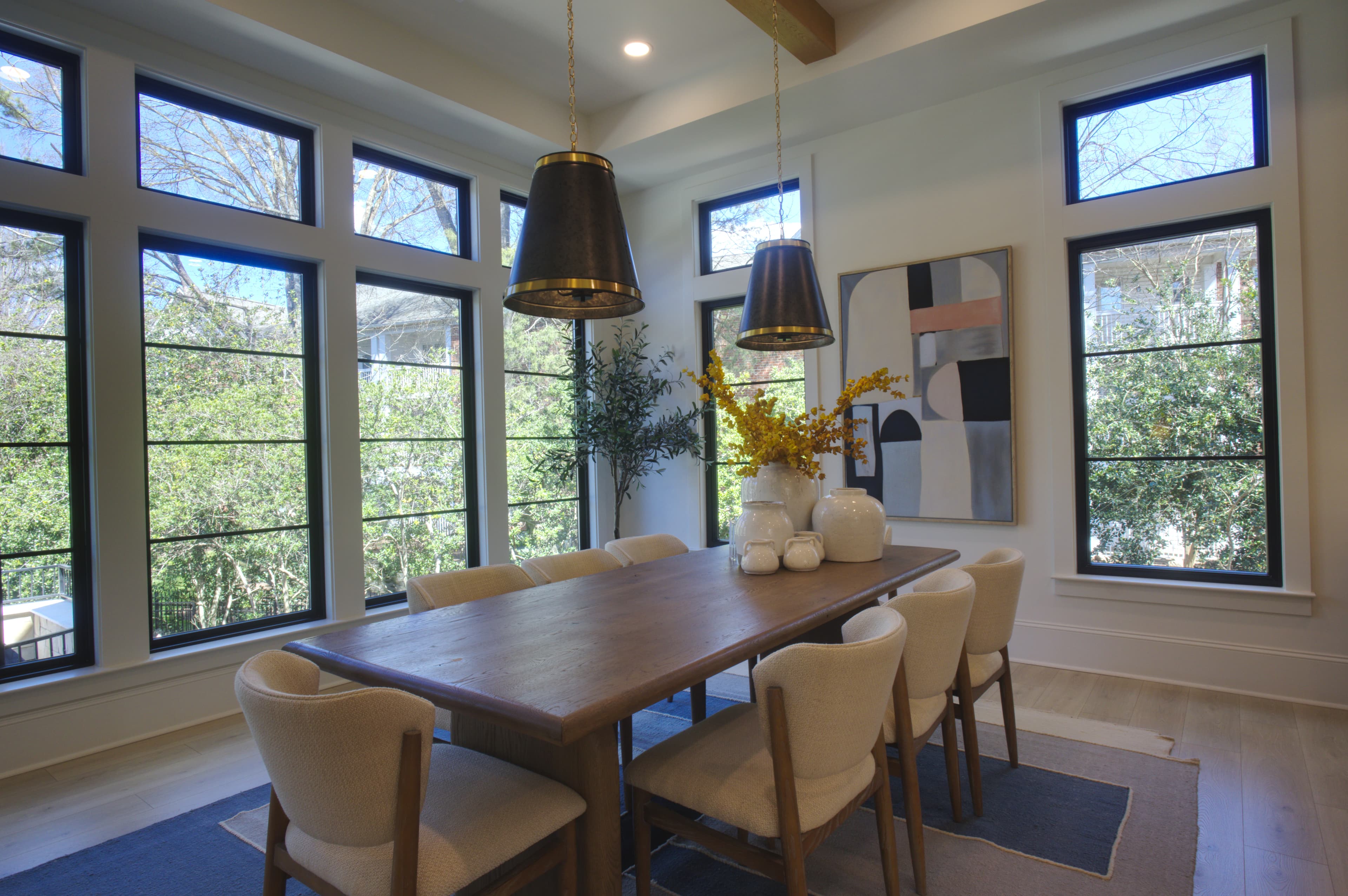 Sunlit modern dining room featuring a wooden table, cream chairs, and large black-framed windows.