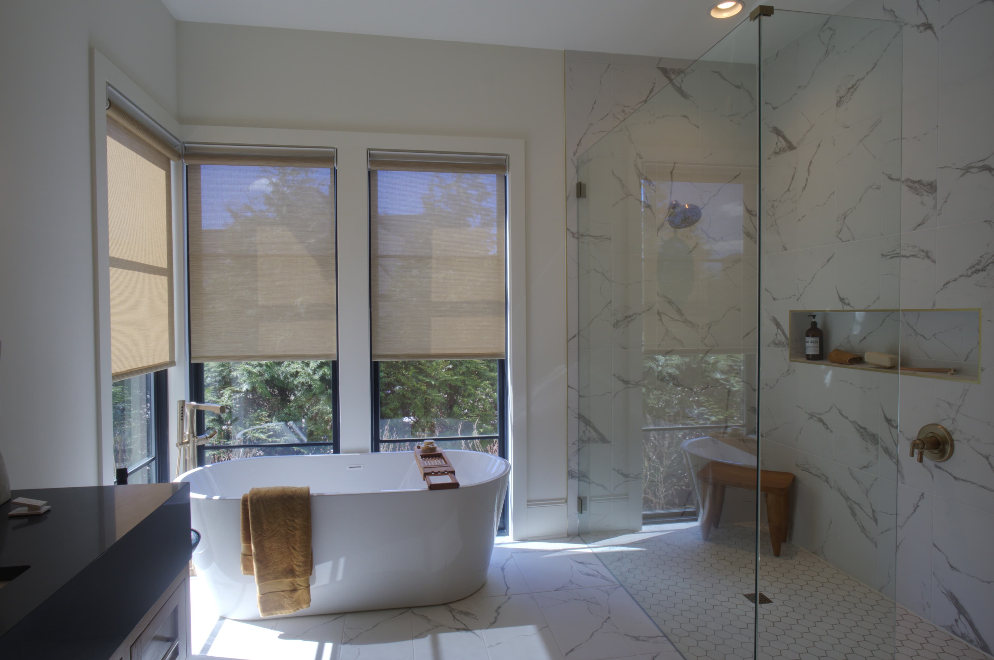 Modern bathroom with a white freestanding tub, marble shower, and large windows with shades.