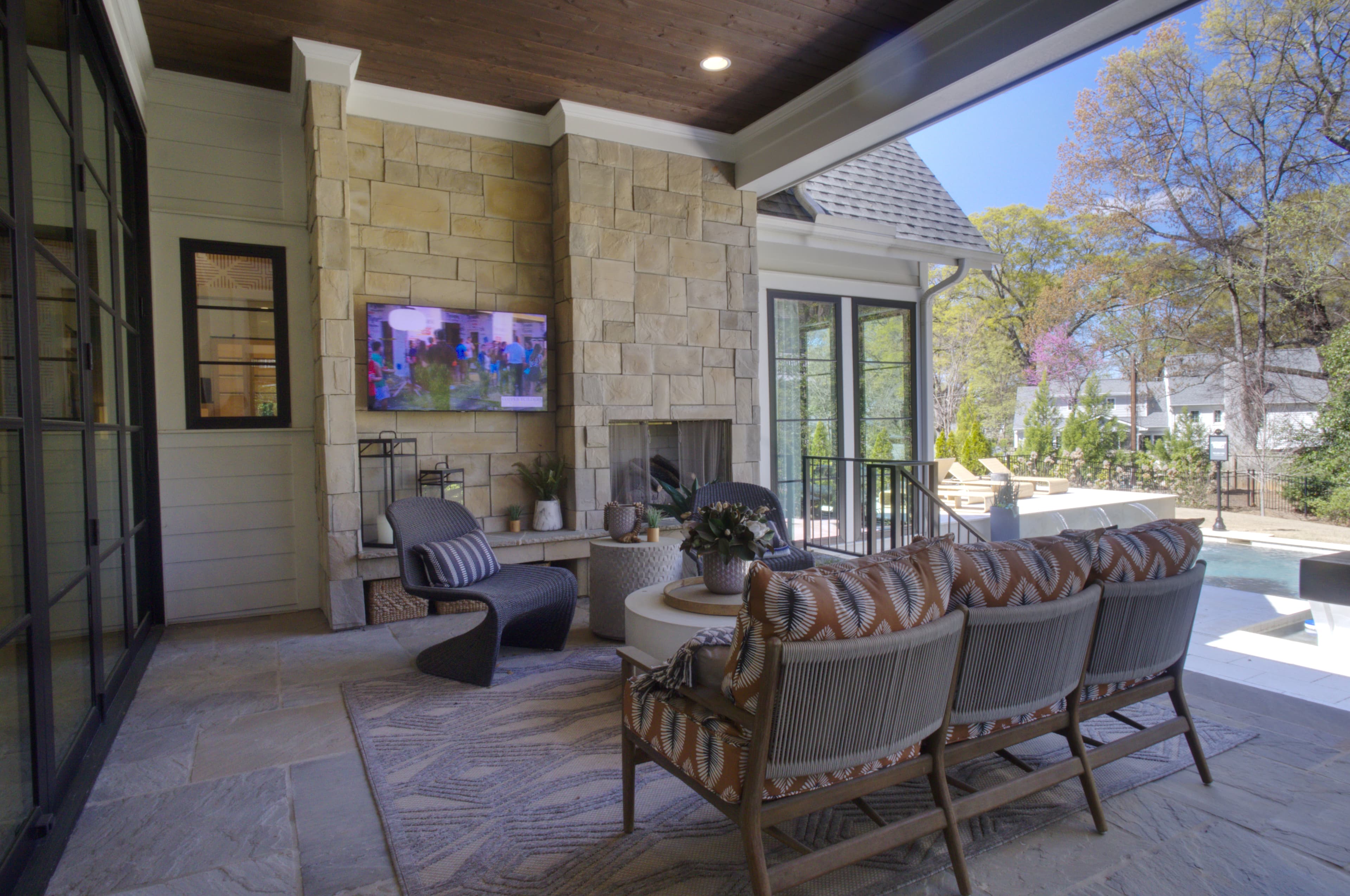 Covered patio with stone fireplace, TV, and modern furniture overlooking a backyard swimming pool.